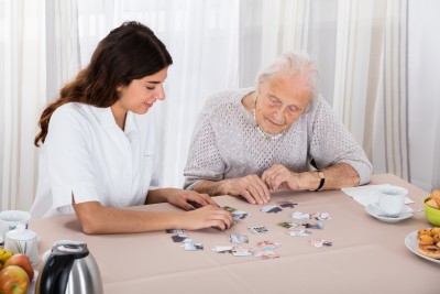 Senior Woman Playing Jigsaw Puzzle On Table With Her Nurse